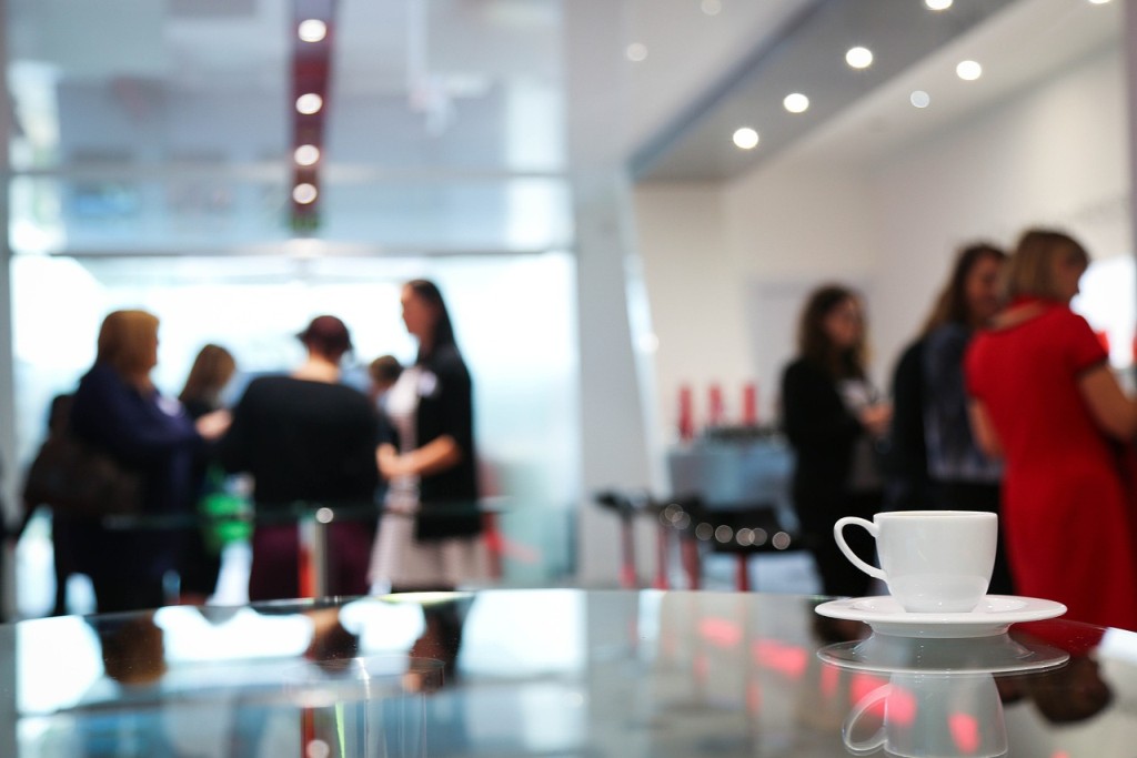 Im Vordergrund steht ein Tisch mit einer Kaffeetasse. Im Hintergrund unterhalten sich mehrere Personengruppen.